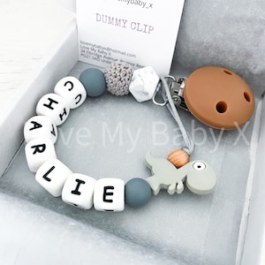 May include: A white silicone baby teether with the name "CHARLIE" spelled out in black letters. The teether is attached to a brown silicone pacifier clip with a gray dinosaur charm and a gray and white marble bead.