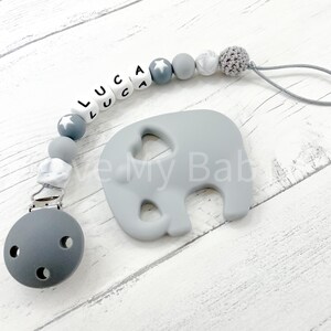 May include: Grey and white silicone baby teether with a grey elephant shape and the name "LUCA" spelled out in white beads. The teether is attached to a grey clip.