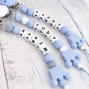May include: Three baby dummy clips with names spelled out in white letter beads. The clips are light blue, white, and grey, with a crown-shaped charm. The names are Paddy, Cody, and Karim. The text "LOVE MY BABY X" is in the lower left corner.