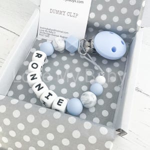 May include: A baby pacifier clip with the name "RONNIE" spelled out in white letter beads. The clip features blue, white, and marble-patterned beads, a blue pacifier, and a metal clip. The item is presented in a white box with a gray polka dot lining.