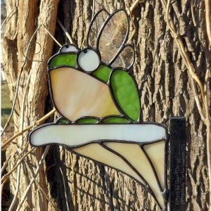 May include: A stained glass frog with wings perched on a mushroom-shaped base. The frog is green and white with large, round eyes. The base is white with a brown border. The frog is a whimsical and colorful piece of art.