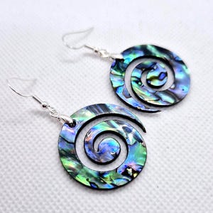 May include: A pair of drop earrings featuring circular abalone shell pendants with a spiral cutout design. The iridescent shell displays vibrant shades of blue, green, and purple. Each pendant is attached to a silver-toned metal fishhook ear wire.