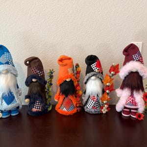 May include: A collection of six decorative gnomes with long white beards and tall hats. The gnomes are dressed in various colors, including blue, brown, orange, black, and burgundy, with plaid accents. Each gnome is adorned with floral or leaf accents.