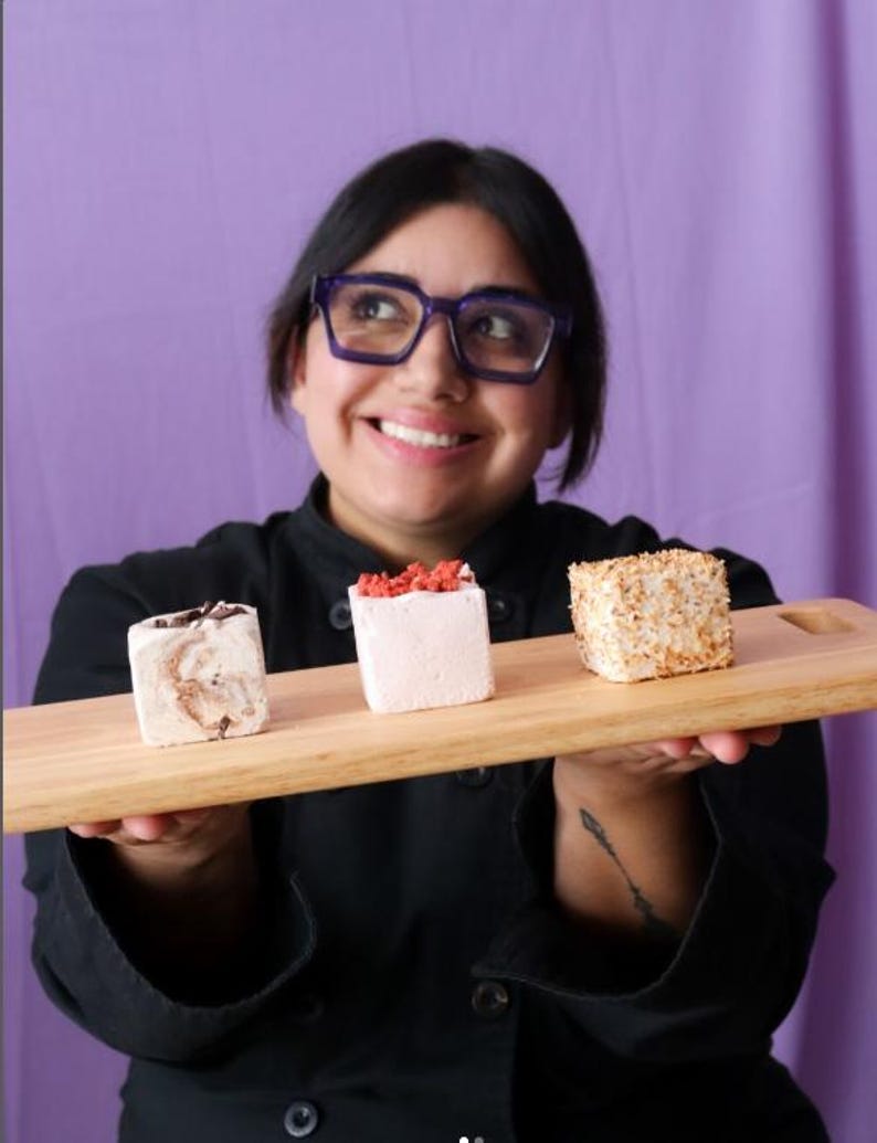 Puede incluir: Una persona sosteniendo una tabla de madera con tres malvaviscos de diferentes sabores. Los malvaviscos est&aacute;n cubiertos con chocolate, chispas rojas y semillas de s&eacute;samo blancas.
