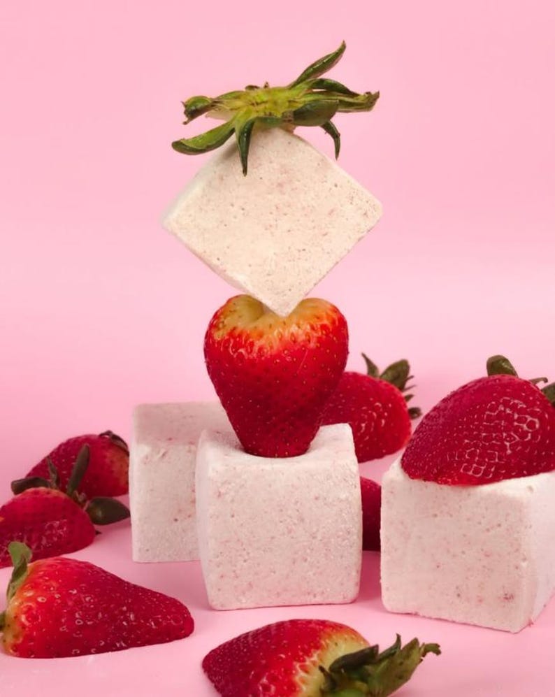 Puede incluir: Malvaviscos cuadrados de color rosa y blanco apilados uno encima del otro con una fresa encima del segundo malvavisco. Hay varias fresas esparcidas alrededor de los malvaviscos sobre un fondo rosa.
