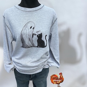 May include: Light gray long-sleeve shirt with a crew neck and a graphic of a ghost and a black cat. The ghost is offering something to the cat. The shirt has dark gray stitching.