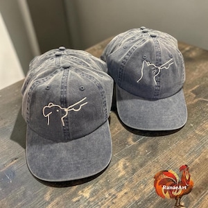 May include: Two charcoal gray baseball caps with a distressed finish. Each cap features a simple white line art embroidery on the front panel. One cap displays a dog silhouette, while the other cap displays a cat silhouette. Both caps have a curved brim.