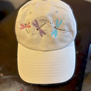May include: A beige baseball cap embroidered with three dragonflies in pink, purple, and blue. The cap has a curved brim and a button on top. The dragonflies are arranged in a row across the front of the cap.