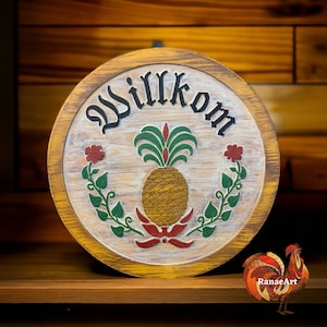 May include: A round wooden sign with the word "Willkommen" painted in black lettering. The sign is decorated with a painted pineapple, green leaves, and red flowers.