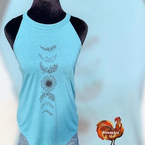 May include: A light blue tank top with a floral moon phase design. The design includes a sunflower and crescent moon shapes. The tank top is displayed on a black mannequin. The background is a light blue color.