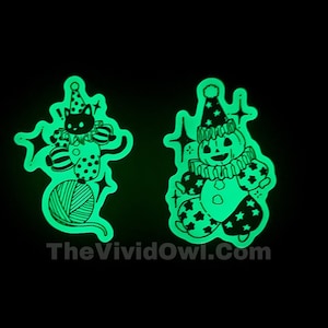 May include: Two glow-in-the-dark stickers featuring a black cat dressed as a clown juggling yarn balls and a pumpkin dressed as a clown. The stickers are green and black and have a whimsical, spooky aesthetic.