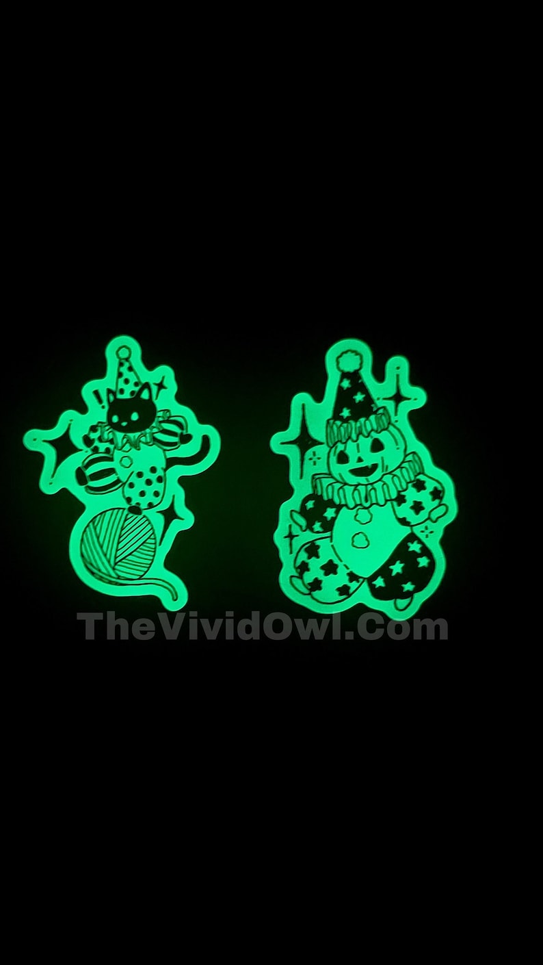 Two Glow in the dark vinyl stickers. One is a spooky Pumpkin dresses as a circus performer. The other is a spooky Black Cat dressed as a circus Performer, balancing on top of a ball of yarn. Set against a dark background with an eerie greenish glow