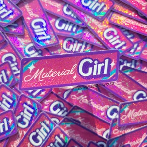 May include: A pile of pink and purple stickers with the words "Material Girl" and "Girl" in white script. The stickers have a glittery, holographic effect and a rectangular shape. The Etsy shop name is visible.