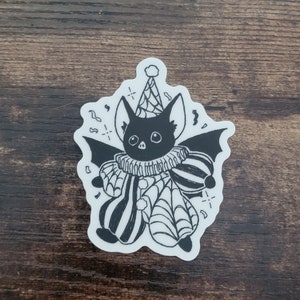 May include: Black and white sticker of a bat wearing a party hat and a jester's costume with a spiderweb pattern. The bat is surrounded by confetti.