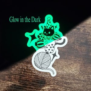 May include: A glow-in-the-dark sticker of a black cat wearing a party hat and holding a ball of yarn. The sticker is on a wooden surface. "Glow in the Dark" is written in green text at the top of the image.