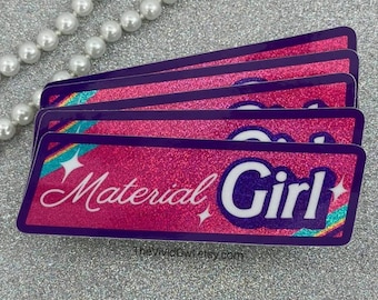 May include: A stack of four rectangular stickers with a pink glitter background and a purple border. The stickers have the words "Material Girl" in white script and a white sans-serif font. The stickers have a rainbow and star design.