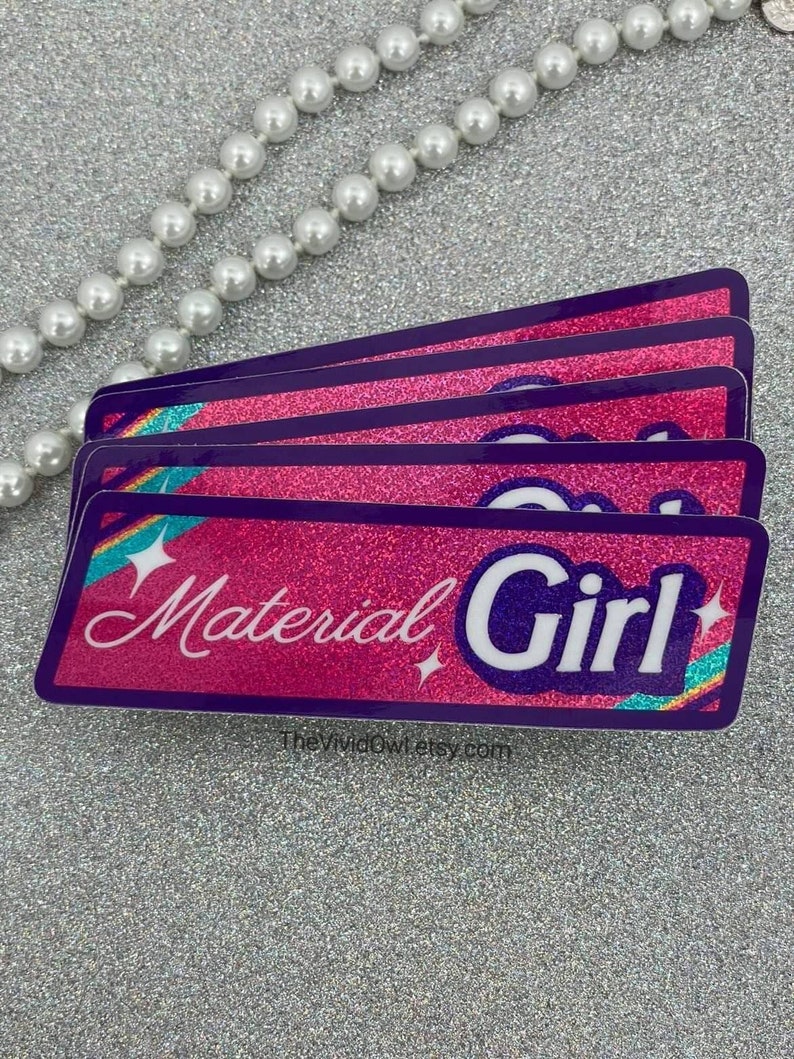 May include: A stack of four rectangular stickers with a pink glitter background and a purple border. The stickers have the words "Material Girl" in white script and a white sans-serif font. The stickers have a rainbow and star design.