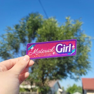 May include: A rectangular sticker with the words "Material Girl" in white and blue, set against a pink glitter background. The sticker features a rainbow and stars design. The VividOwl.etsy.com is printed at the bottom.