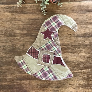 May include: A fabric witch hat decoration featuring a patchwork design of plaid and solid tan, maroon, and green fabric segments. The hat includes a central maroon star applique and a rectangular buckle detail, all finished with visible white stitching.