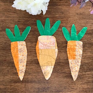May include: Three handmade fabric carrots with green tops and orange and yellow bodies. The carrot bodies have a patchwork design with floral patterns. The carrots are arranged on a wooden surface.