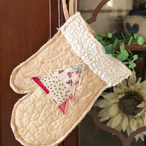 May include: A quilted beige Christmas stocking with a white sherpa cuff and a fabric Christmas tree applique. The tree is made of patterned fabric with red and floral accents. A loop is attached for hanging.