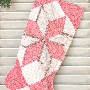 May include: A pink and white patchwork Christmas stocking with a star design. The stocking is made of quilted fabric and has a frayed edge.