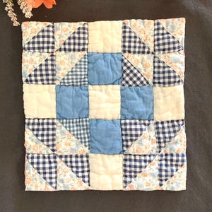 May include: A square quilted fabric piece featuring a patchwork design of blue and white gingham, solid light blue squares, solid white squares, and a floral print fabric with small orange and blue flowers arranged in a grid pattern.