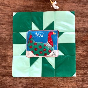 May include: A square fabric quilt block featuring a star pattern in alternating shades of dark and light green. Centered on the block is a rectangular patch depicting a goose with a red neck and green body, with the word Noel written above.