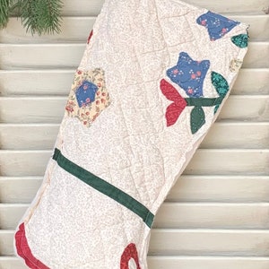 May include: A white quilted Christmas stocking with a red, green, and blue floral design. The stocking has a red bow at the toe.