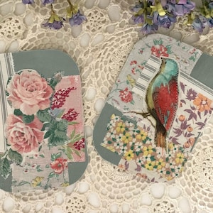 May include: Two decorative, oval-shaped coasters. One features pink roses and floral patterns, the other a colorful bird perched on a branch. Both have a gray border and are set against a lace background.