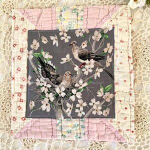 May include: A square, handmade quilt featuring a central panel with a dove and floral print. The dove print is framed by a pink and floral patterned border. The quilt is likely made of cotton and has a vintage aesthetic.
