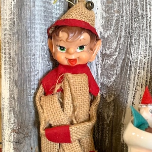 May include: A vintage Christmas elf ornament with a burlap body and red accents. The elf has a painted face with green eyes, a red hat, and a burlap hat. The ornament is displayed on a rustic wooden surface.