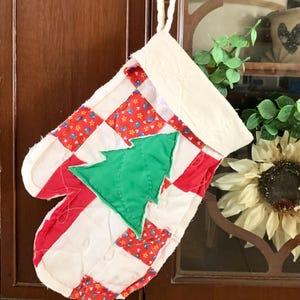 May include: A quilted Christmas stocking shaped like an oven mitt. The stocking features a patchwork design with red and white stripes and squares, and a green Christmas tree applique. It hangs from a white cord.