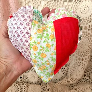 May include: A patchwork heart-shaped pillow made from different floral and patterned fabrics. The pillow is red, white, green, and purple.
