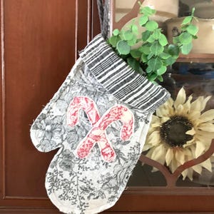 May include: A quilted oven mitt with a floral pattern in gray and white, featuring two red and white candy cane appliqués. The top is striped in black and white, and it hangs from a gray rope. Greenery is visible inside the mitt.
