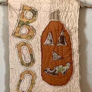 May include: A quilted Halloween wall hanging with an orange pumpkin and the word "BOO" in appliqued letters. The pumpkin has triangle eyes and a jagged mouth. The background is off-white with a stitched pattern.