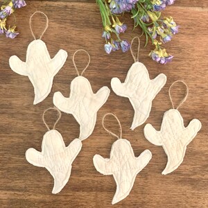 May include: Six white fabric ghost ornaments with stitched details and jute twine hangers are arranged on a wooden surface. Purple flowers are in the background. These Halloween decorations are perfect for adding a spooky touch to any space.