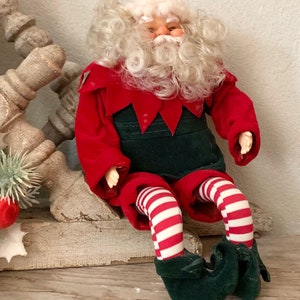 May include: A plush Santa Claus doll with a white beard and curly white hair, wearing a red and green elf costume with white and red striped stockings and green shoes. The doll is sitting on a wooden surface.