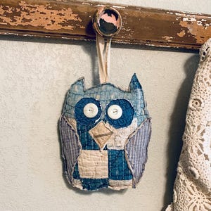 May include: A handcrafted owl ornament made from quilted fabric patches in shades of blue, white, and grey. The owl features two white button eyes, a triangular fabric beak, and a fabric loop at the top for hanging, showcasing visible stitching.