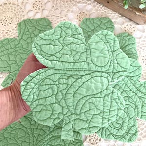 May include: A collection of light green, quilted shamrock-shaped coasters. The coasters have a detailed, stitched pattern and a soft, textured appearance. The shamrocks are arranged on a cream-colored, lace-like surface.