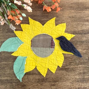 May include: A quilted fabric sunflower applique featuring bright yellow petals with stitched grid patterns, a brown plaid circular center, two light green leaves, and a dark navy blue bird silhouette perched on the right side of the flower.