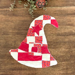 May include: A quilted fabric witch hat ornament featuring a patchwork design of red, white, and floral patterned squares. The hat includes a red star appliqué on the crown and a square buckle detail on the brim, with visible decorative stitching.
