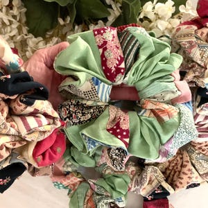 May include: A collection of colorful scrunchies in various patterns and colors. The scrunchies feature a mix of fabrics, including green satin, floral prints, and striped designs. The scrunchies are arranged together, with a background of white flowers and green leaves.