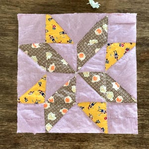May include: A square quilt block featuring a pinwheel pattern made of fabric patches. The design includes light purple gingham, yellow fabric with small figures, and brown fabric with a repeating white and orange floral-like pattern.