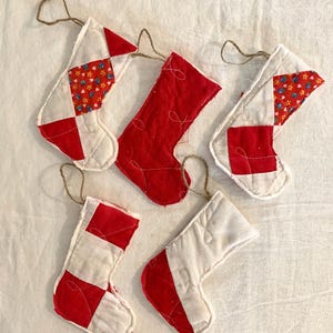May include: Five small, handmade Christmas stockings in red and white patchwork designs. Some stockings feature floral fabric accents. Each stocking has a loop of twine for hanging. The stockings are arranged on a white surface.
