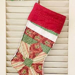May include: A red quilted Christmas stocking with a patchwork design featuring green, pink, and white fabric. The stocking is hanging on a white shutter.