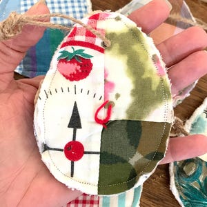 May include: Handmade fabric ornaments in various patterns and colors. One ornament features a compass design with a red dot and arrow, and a strawberry. Each ornament has a twine loop for hanging.