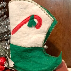 May include: A green and white felt Christmas stocking with a red candy cane and a green holly leaf applique. The stocking has a bell on the side and a pointed toe.