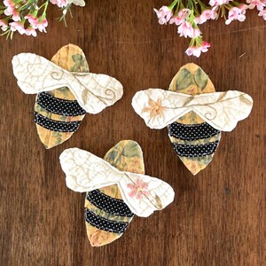 May include: Three handmade bee-shaped fabric ornaments. The bees have cream-colored wings with embroidered details and black and white striped bodies. The background fabric is yellow with a floral pattern. A pink flower is on one wing.