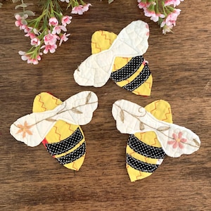May include: Three handmade bee-shaped fabric appliqués. Each bee features yellow and black striped bodies, white wings with embroidered details, and red zigzag stitching. Pink flowers and greenery are in the background on a wooden surface.